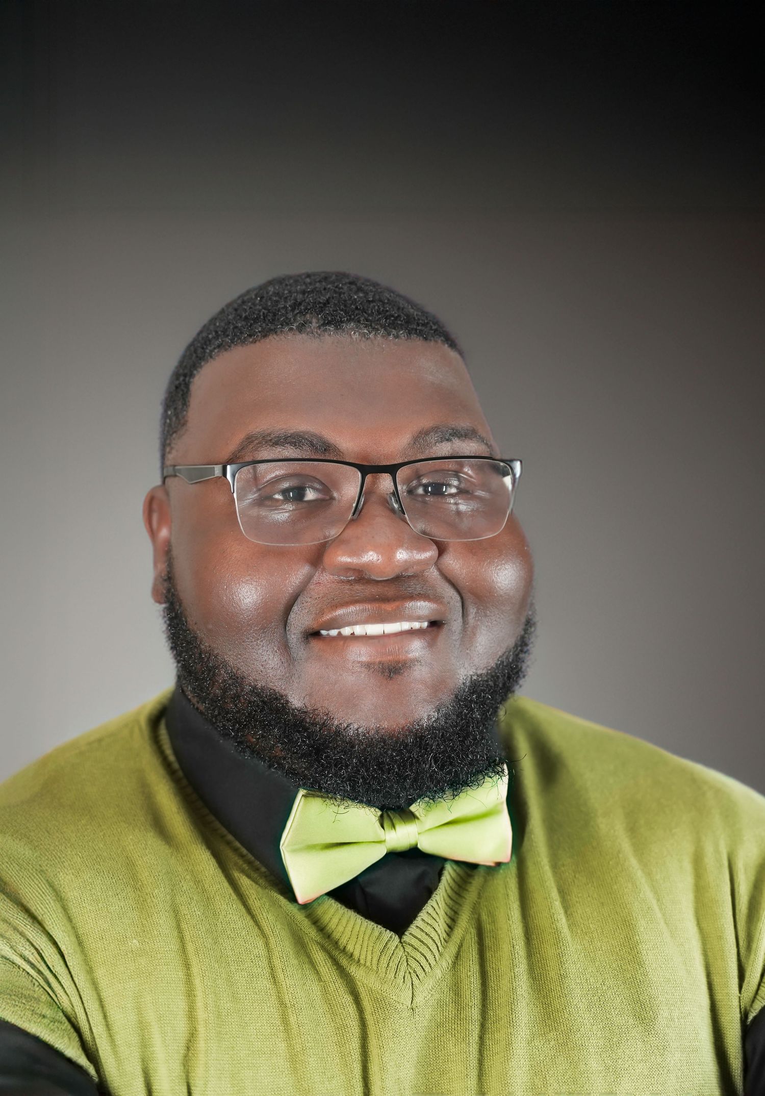 Smiling man with glasses and a green bow tie wearing a green sweater, professional headshot portrait.