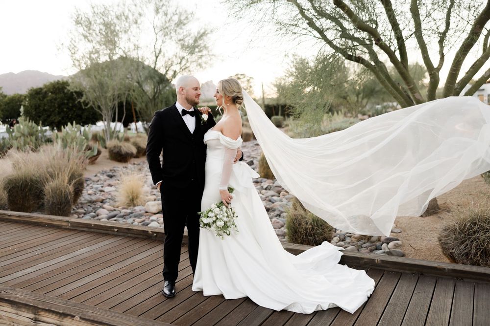 Bride and groom share romantic moment on wooden deck in desert garden, veil flowing in wind, mountains in background.