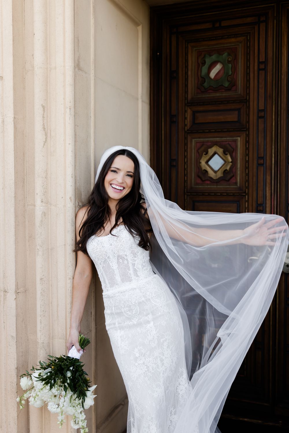 Smiling bride in white lace gown and flowing veil holds bouquet beside ornate wooden door and stone columns.