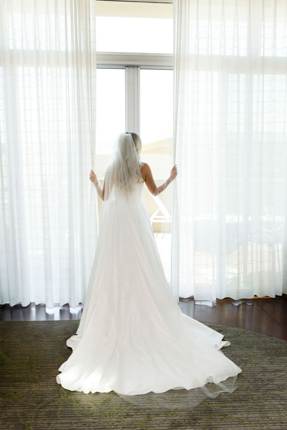 Bride in white wedding gown and veil standing at large window with sheer curtains, viewed from behind.