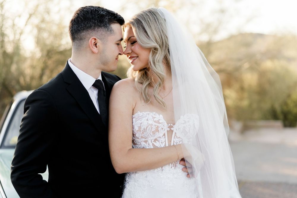 Bride and groom share a romantic moment outdoors, with the bride in a lace gown and veil, groom in a black tuxedo.