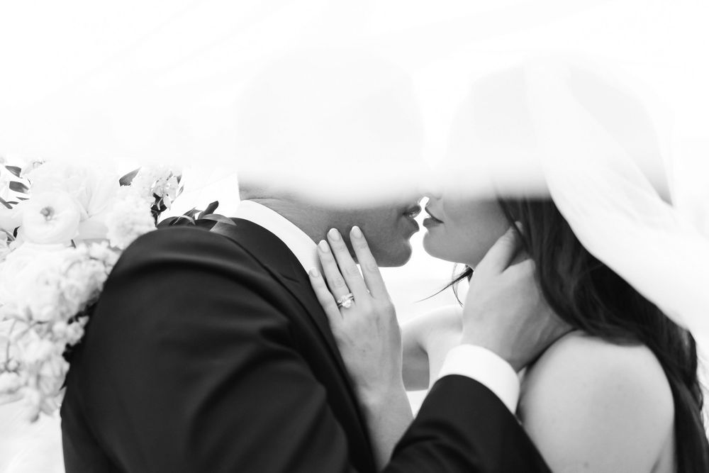 Black and white wedding photo of bride and groom sharing a kiss under veil with flowers nearby.