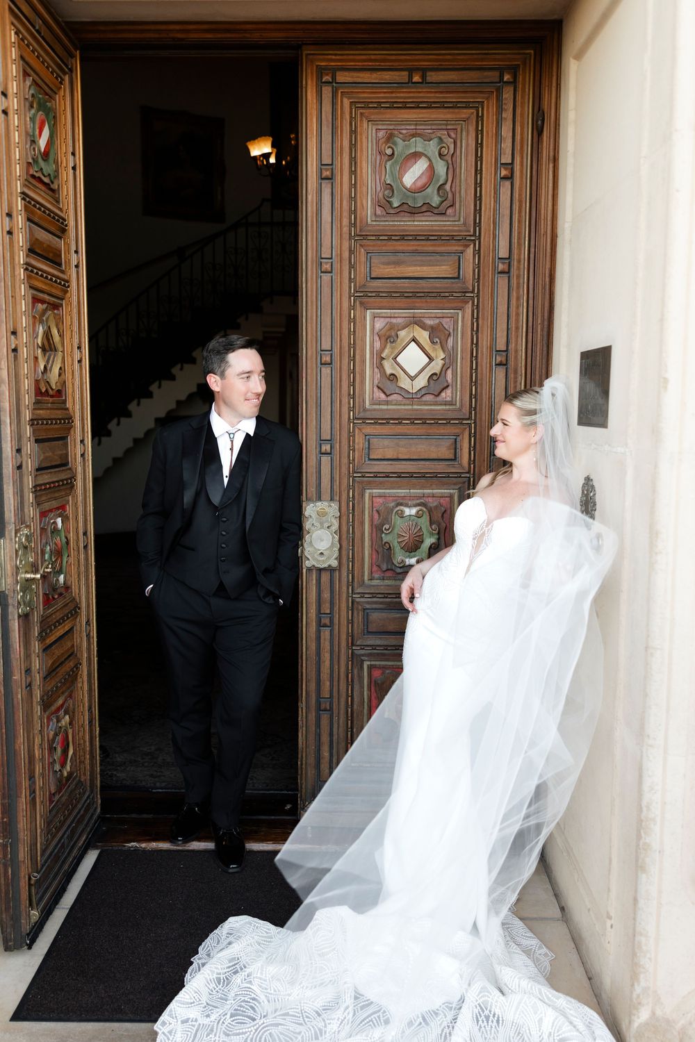 Bride in flowing white gown and veil stands with groom in black suit at ornate wooden church doors.