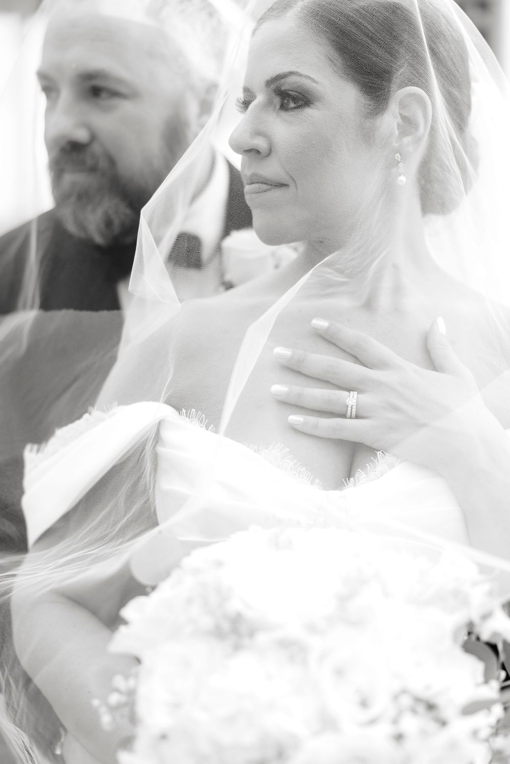 Black and white photo of an emotional bride in veil holding bouquet with bearded man in background.