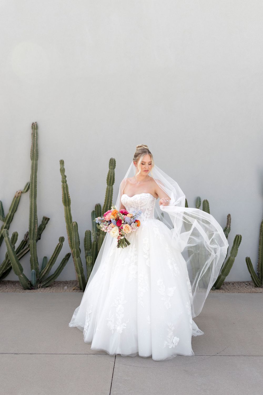 Bride in white ballgown with flowing veil holds colorful bouquet beside tall cacti against a light gray wall.