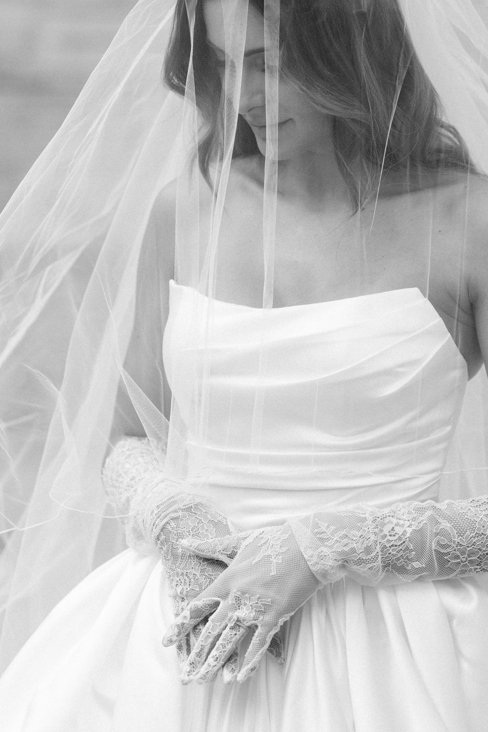 Black and white photo of a bride in a strapless gown with lace gloves, draped in a flowing veil, looking down.