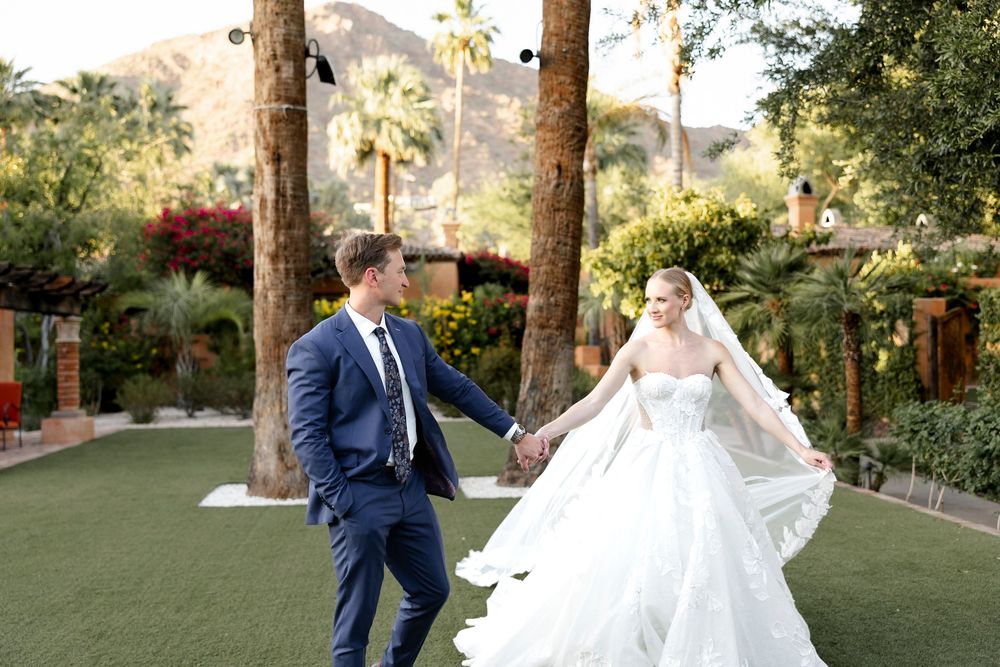 Bride in flowing white gown and groom in navy suit hold hands at an outdoor wedding venue with palm trees.