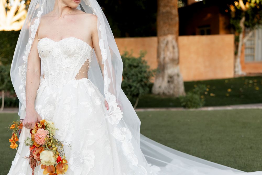 Bride in strapless lace ball gown with cutout detail and veil holding colorful autumn bouquet outdoors.