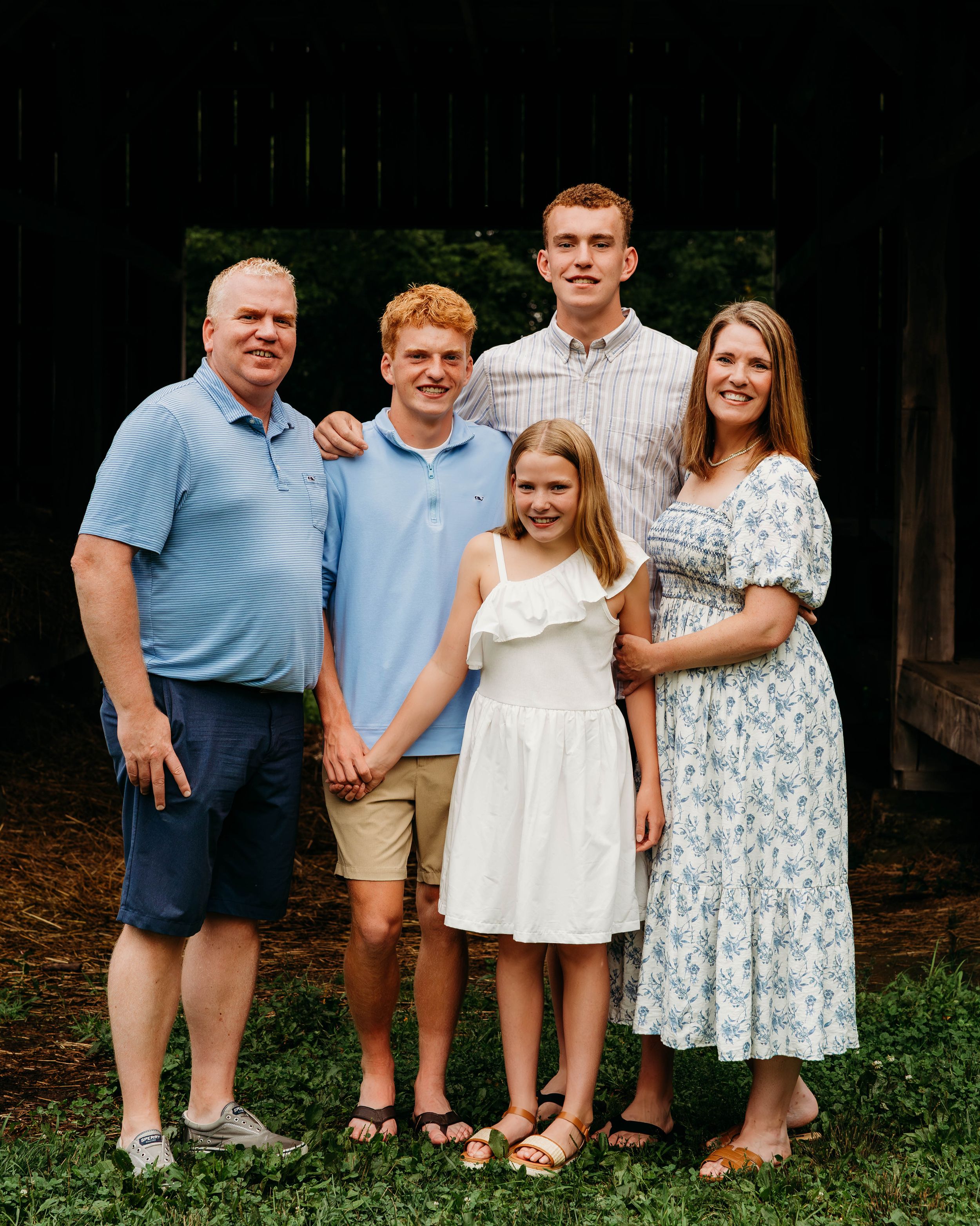 Family of five smiling together outdoors near a rustic barn, wearing coordinated blue and white outfits.