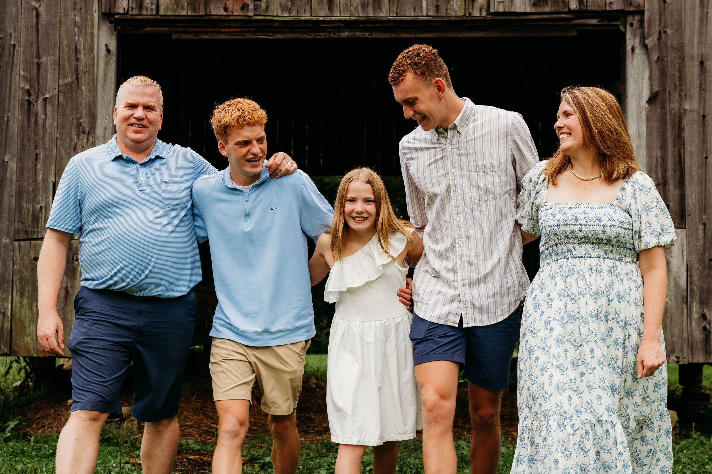 A joyful family of five poses and laughs together outdoors in front of a rustic wooden barn.