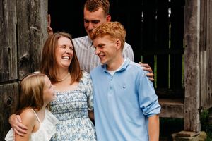 Happy family of four laughing together outdoors near a rustic wooden barn during a casual portrait session.