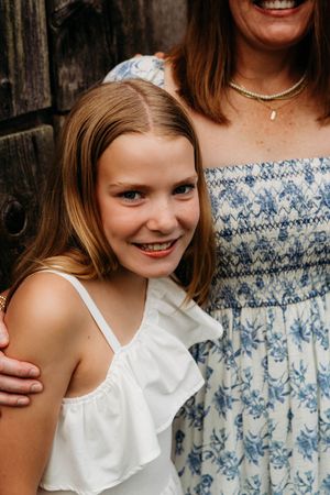 A smiling young girl in a white ruffled top poses with a woman in a blue floral dress outdoors.