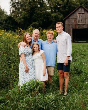 Happy family of five posing in a wildflower field with a rustic barn backdrop on a summer day.