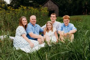 Family of five smiling together in a lush green field with a rustic barn in the background.