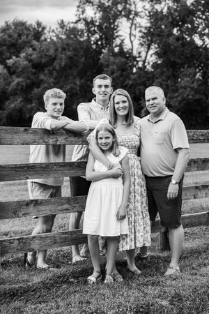 Black and white family portrait of five people posing by a wooden fence outdoors with trees in the background.