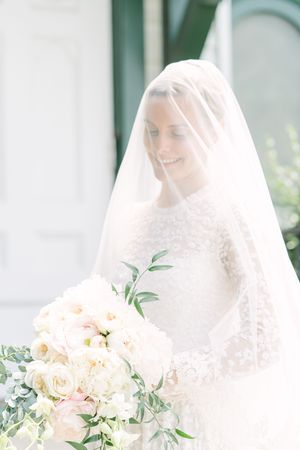 Smiling bride in a lace gown and sheer veil over her face holds a white and blush floral bouquet in soft natural light.