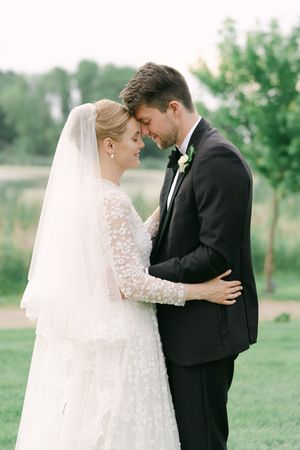 Bride in a lace gown and long veil touches foreheads with her groom in a black tuxedo during an outdoor wedding portrait.