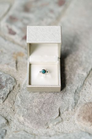 Silver ring with a round teal blue gemstone sits in an open cream ring box on a rough natural stone surface.