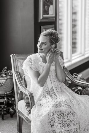 Black and white portrait of a bride in a lace gown seated in an ornate chair by a window, adjusting her earring. Lincoln, NE Photographer