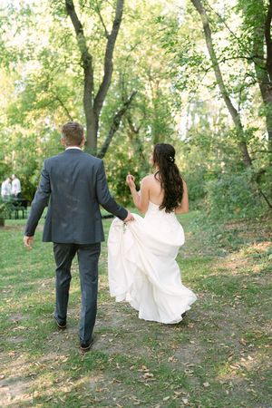 Bride in a flowing white gown and groom in a gray suit walk away from the camera across a sunlit grassy path in a leafy park.