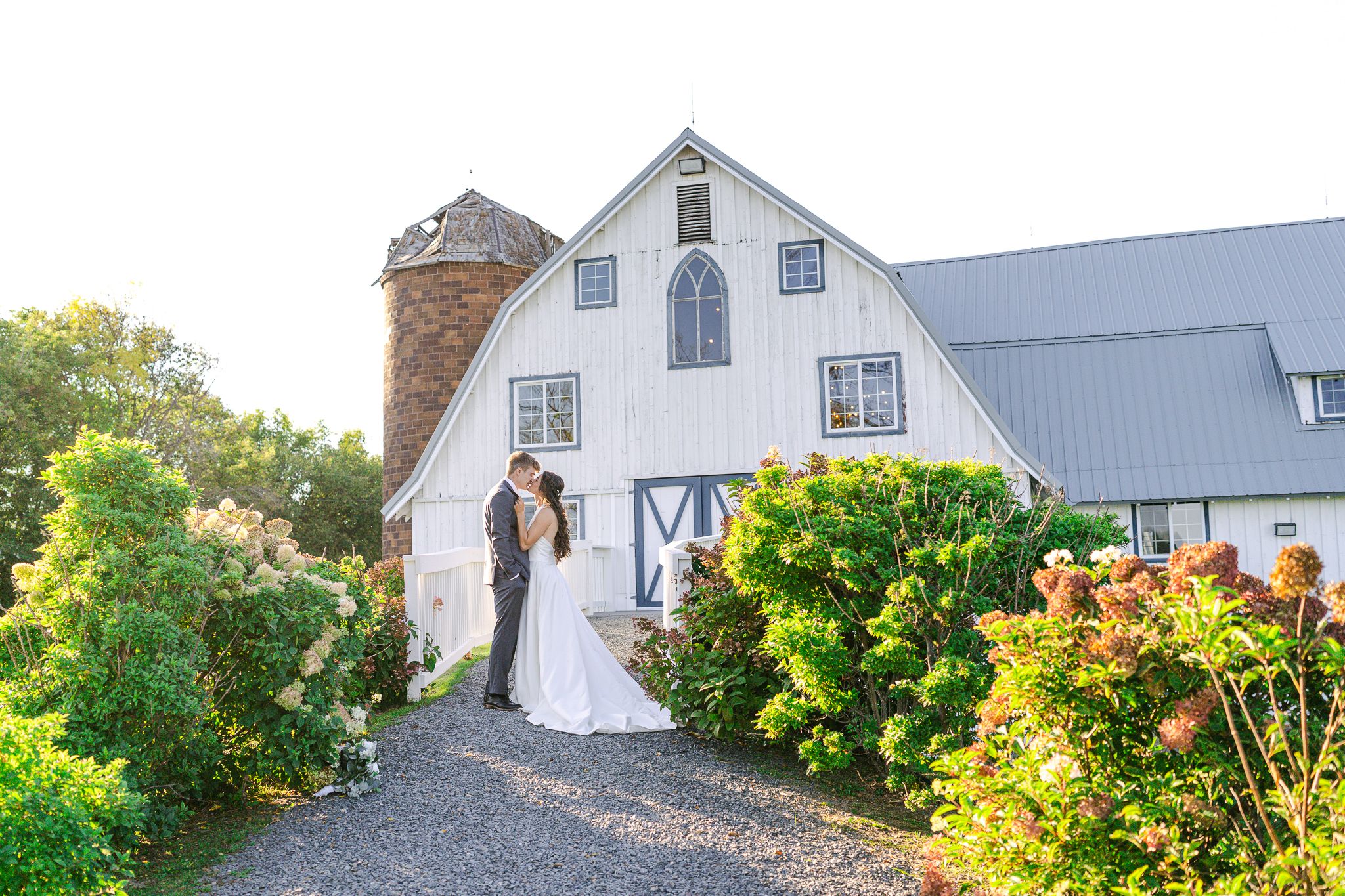 Bride and groom embrace on a gravel path in front of a white barn wedding venue with a stone silo and blooming hydrangeas.