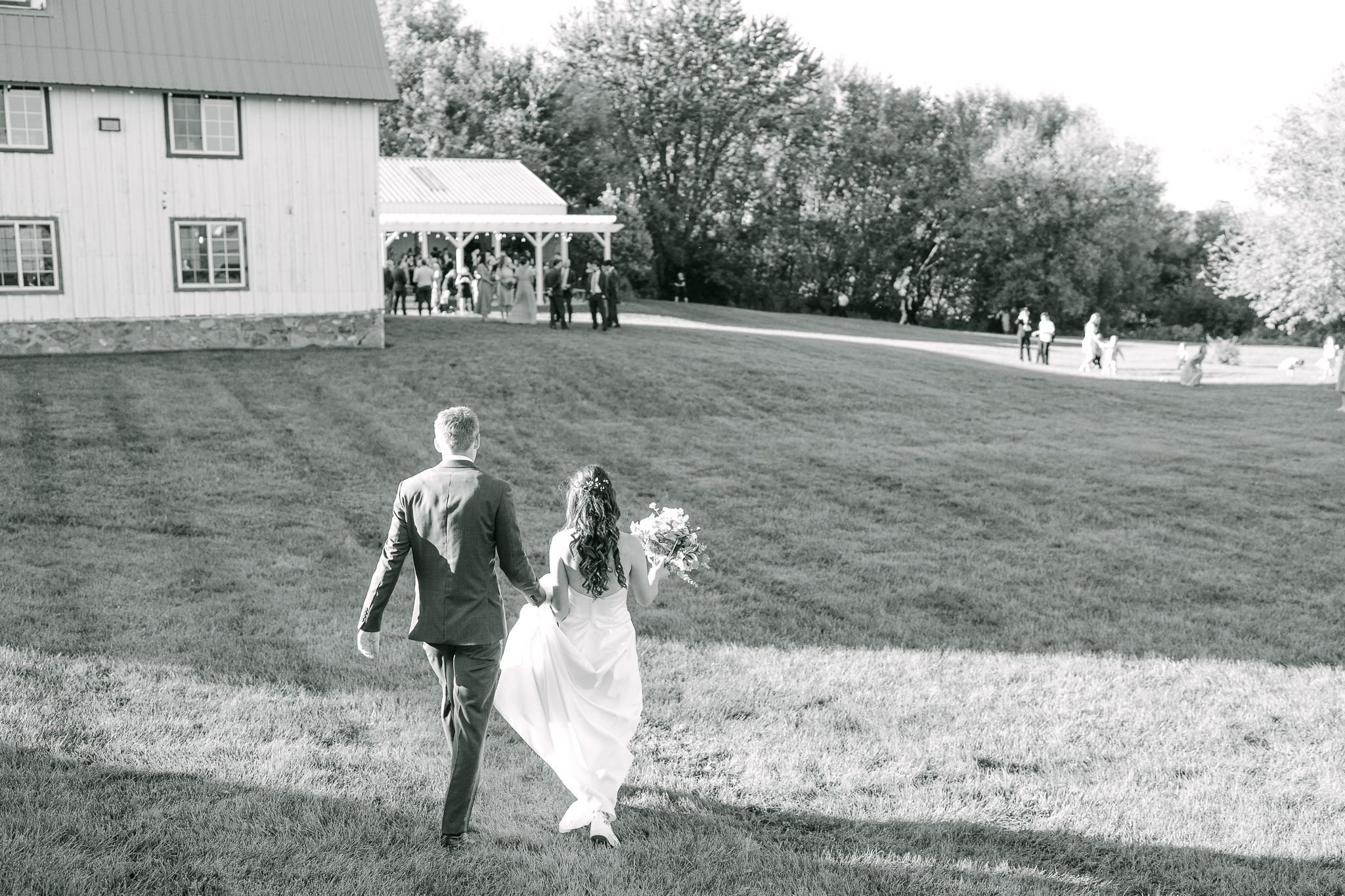 Bride and groom hold hands and walk across a sunlit lawn toward a white pavilion and wedding guests in black and white.