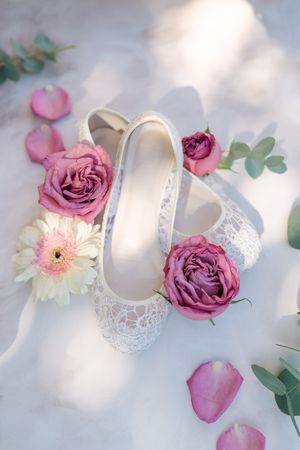 White lace bridal flats surrounded by pink roses, a white gerbera daisy, loose petals and eucalyptus leaves on soft white fabric.