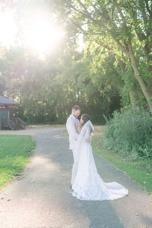 Bride and groom share an intimate moment on a park path at sunset, her long veil and lace wedding gown flowing among sunlit trees.