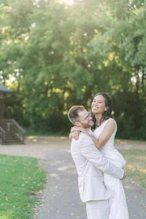 Joyful couple in light wedding attire embracing on a park path, the bride laughing as the groom lifts her, surrounded by sunlit green trees.