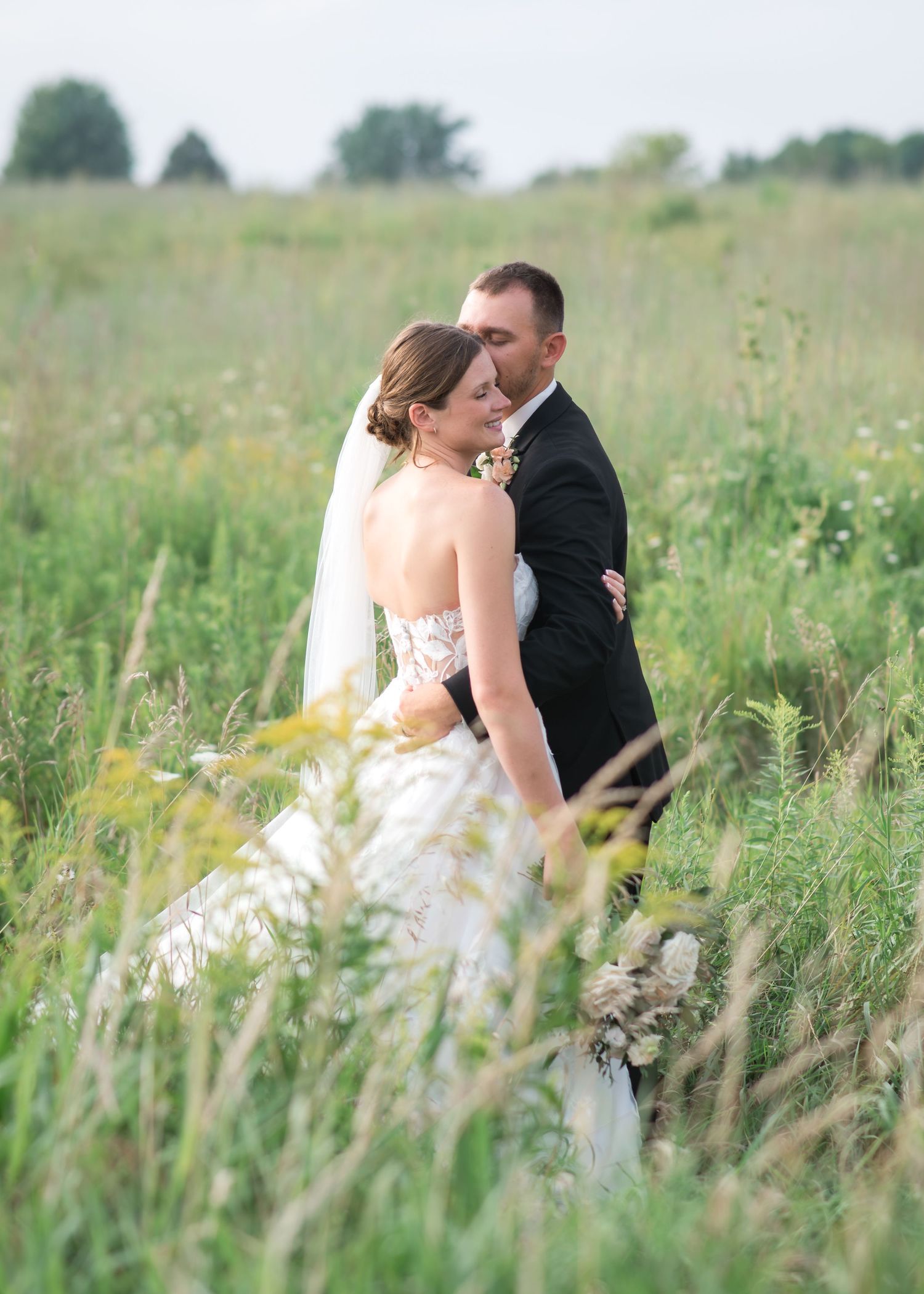 Bride in a lace wedding gown and veil embraces her groom in a dark suit amid tall green grasses in a sunlit meadow.