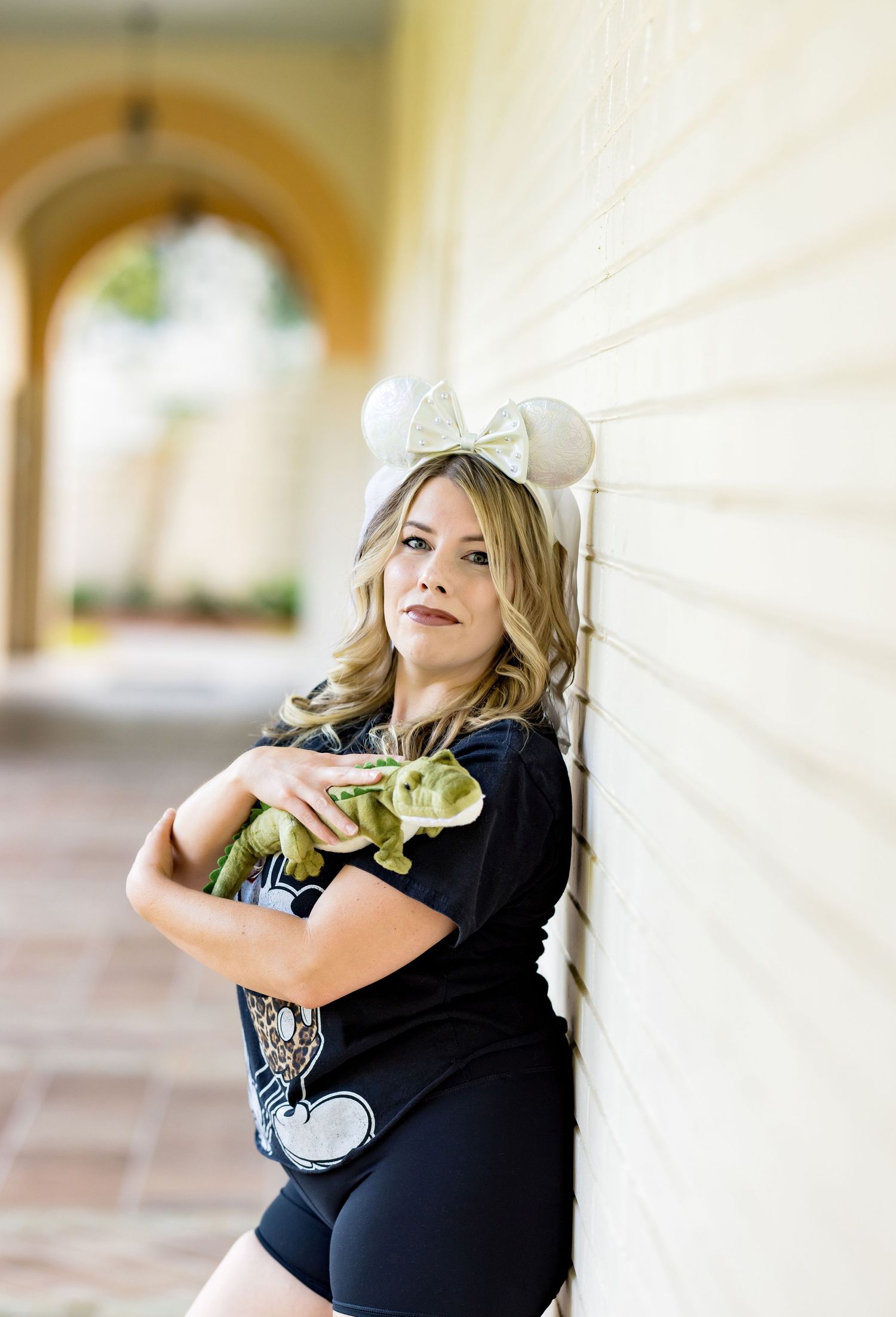 Woman in Mickey Mouse ears holds a green dinosaur plush toy while leaning against a white wall in a sunny arcade.