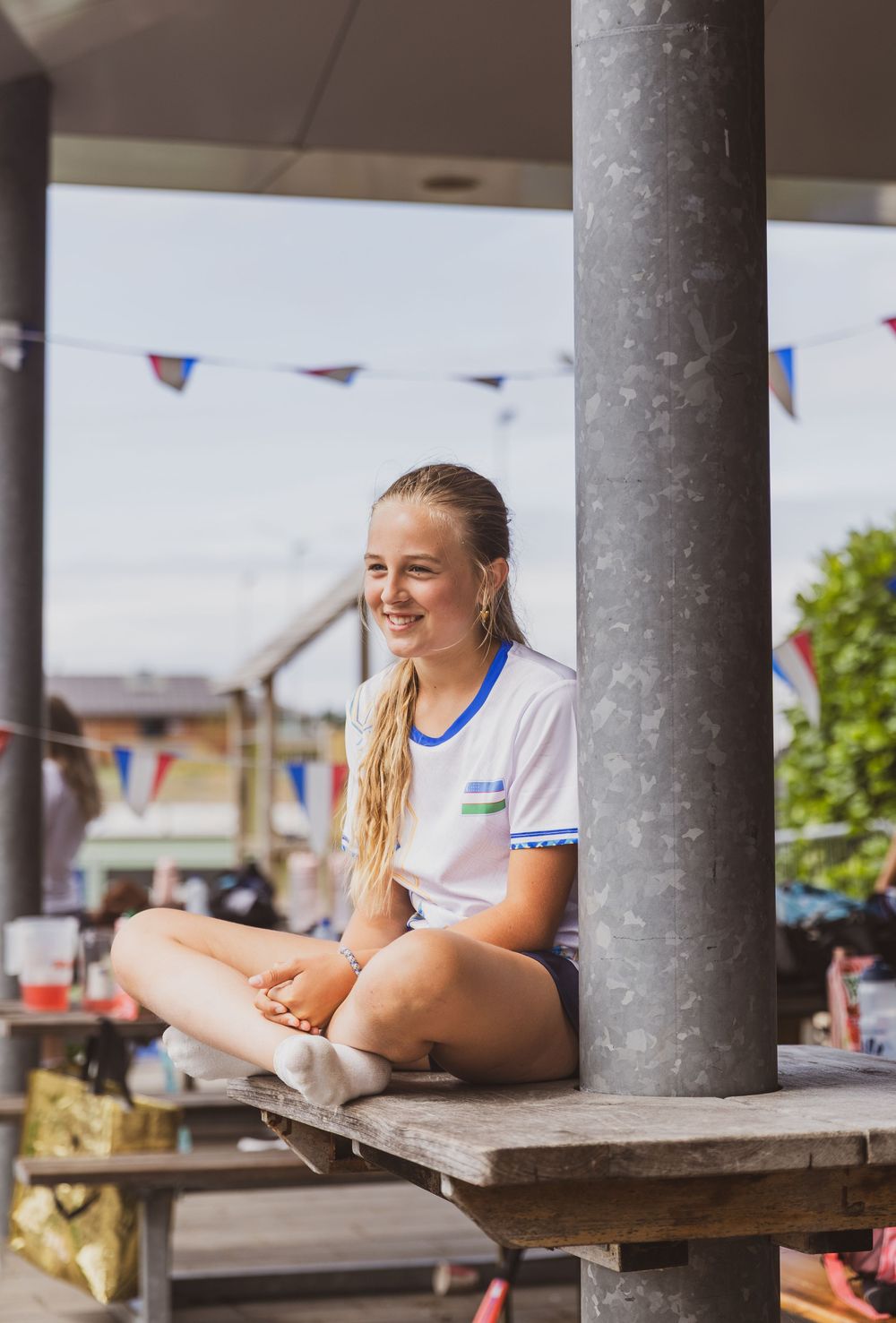 Young woman in white shirt sits cross-legged on wooden table outdoors, smiling, with colorful bunting flags in background.