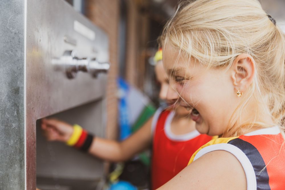 Smiling blonde girl interacting with a metal vending machine outdoors, wearing a colorful tank top.