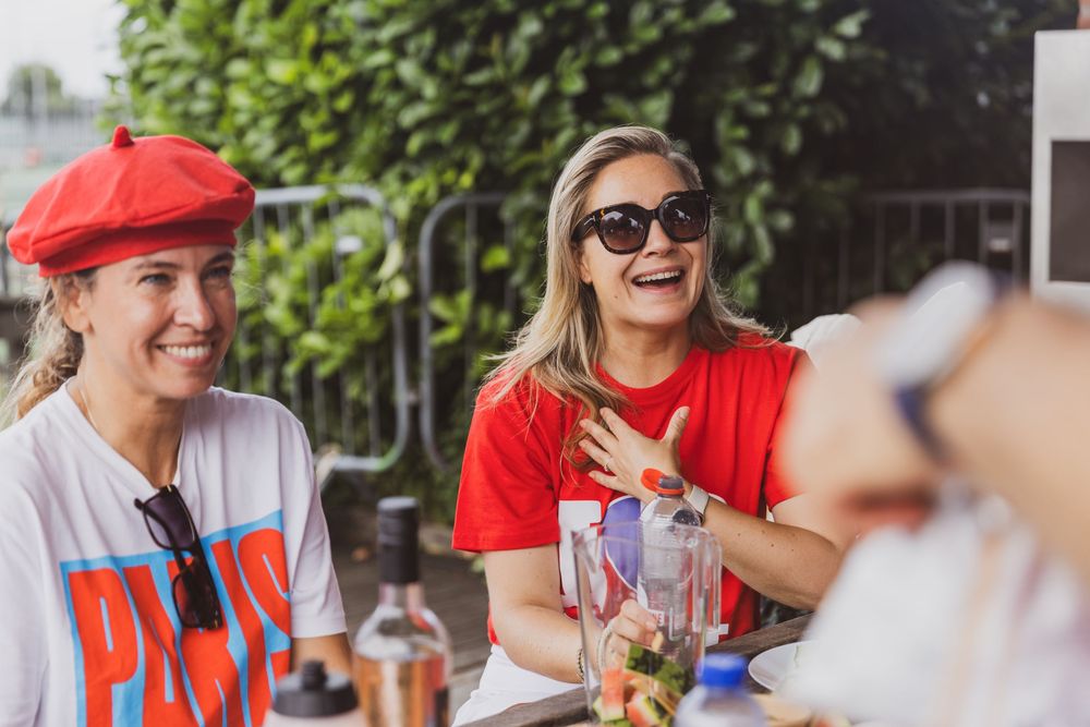 Two women laughing outdoors at a table, one wearing a red beret and Paris shirt, the other in red top and sunglasses.