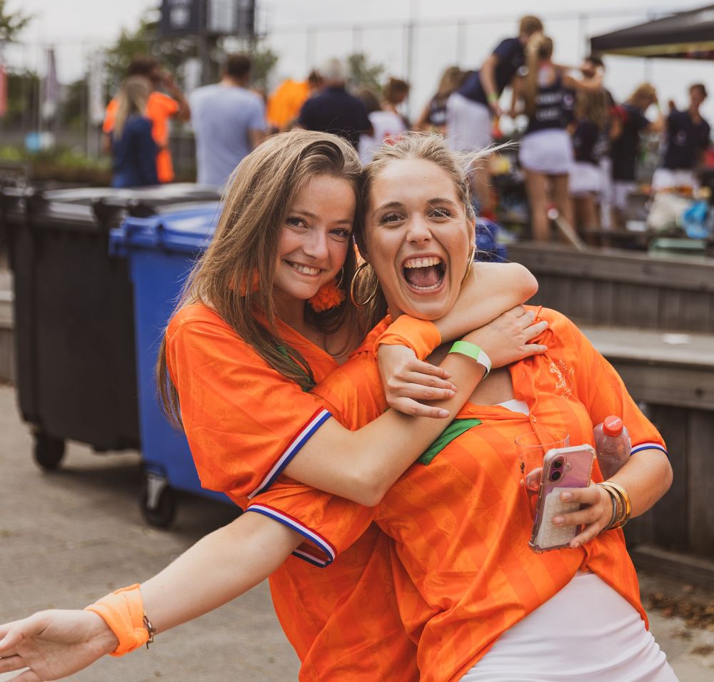 Two young women in orange jerseys hugging and smiling at an outdoor event.