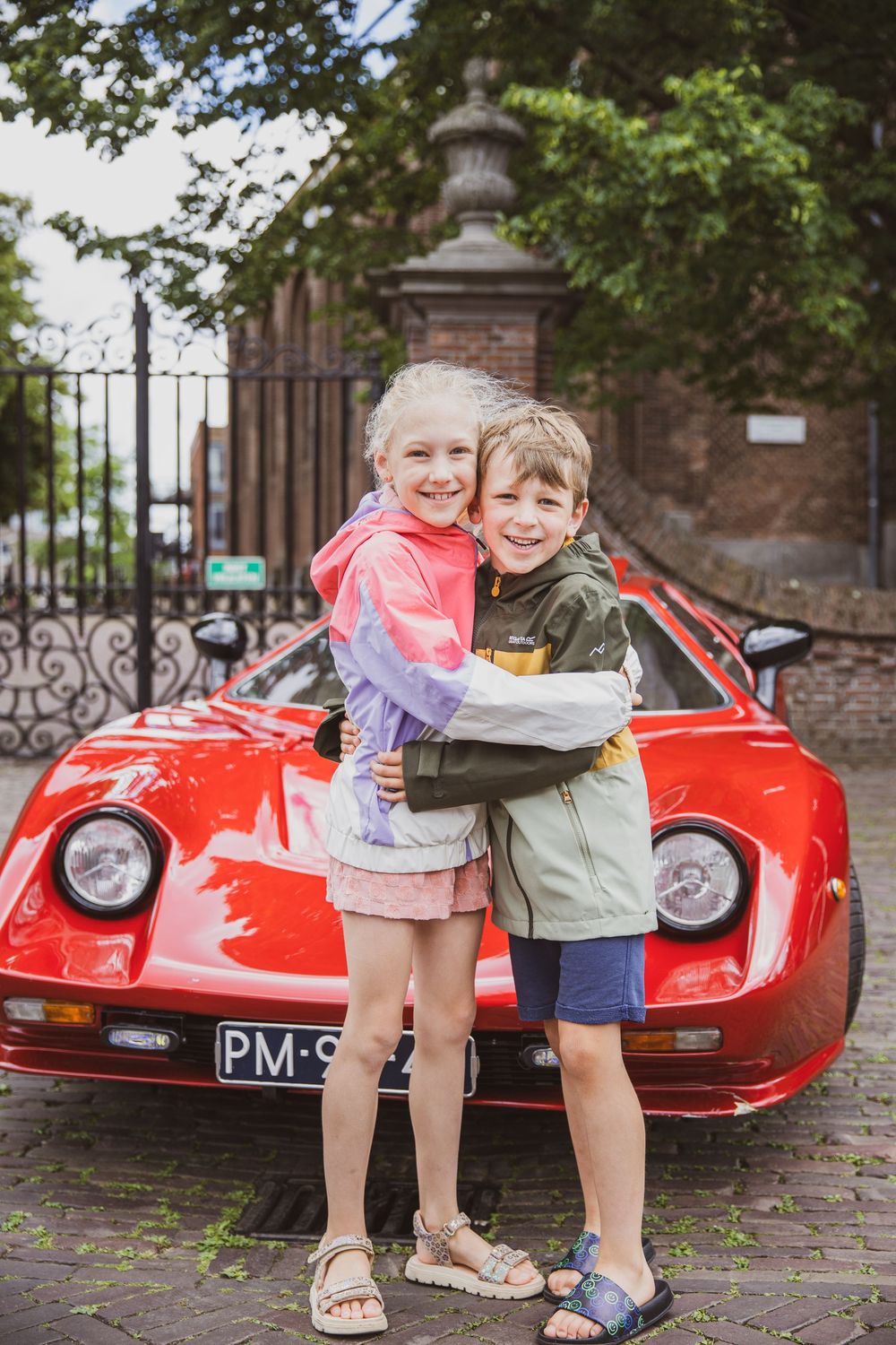 Two children hugging and smiling in front of a classic red Ferrari sports car on a cobblestone street.