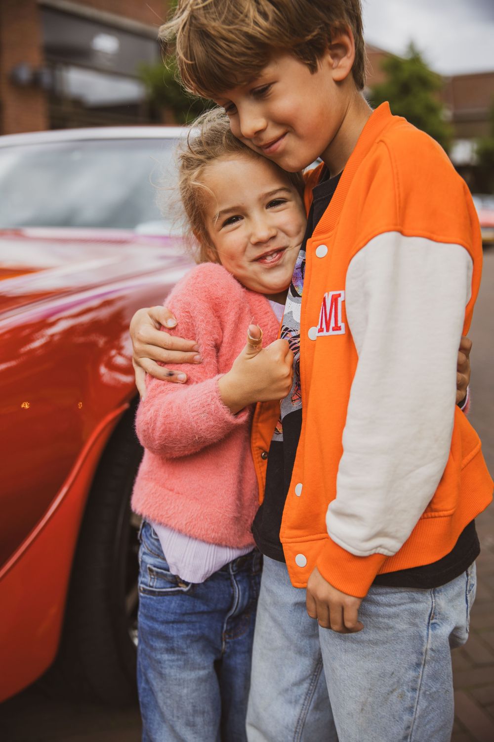 Two children hugging and smiling in front of a red car, wearing colorful jackets and jeans.