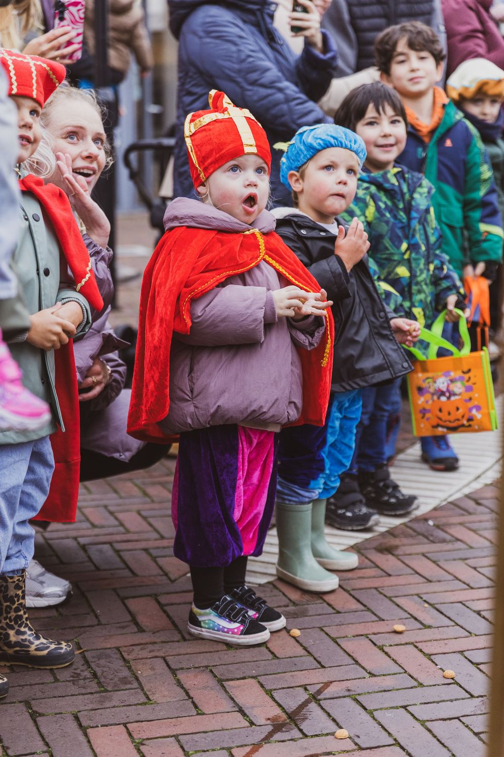 Children in festive costumes watch a Sinterklaas parade on a brick street, with one child dressed as Sinterklaas.