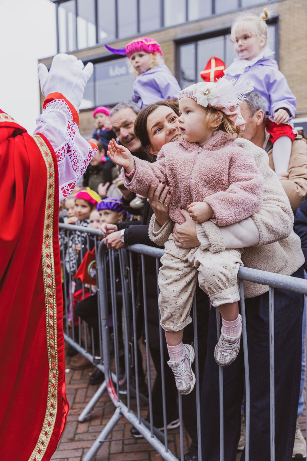 Child reaches toward Sinterklaas in red robe at a festive Dutch holiday parade, with crowd behind metal barriers.