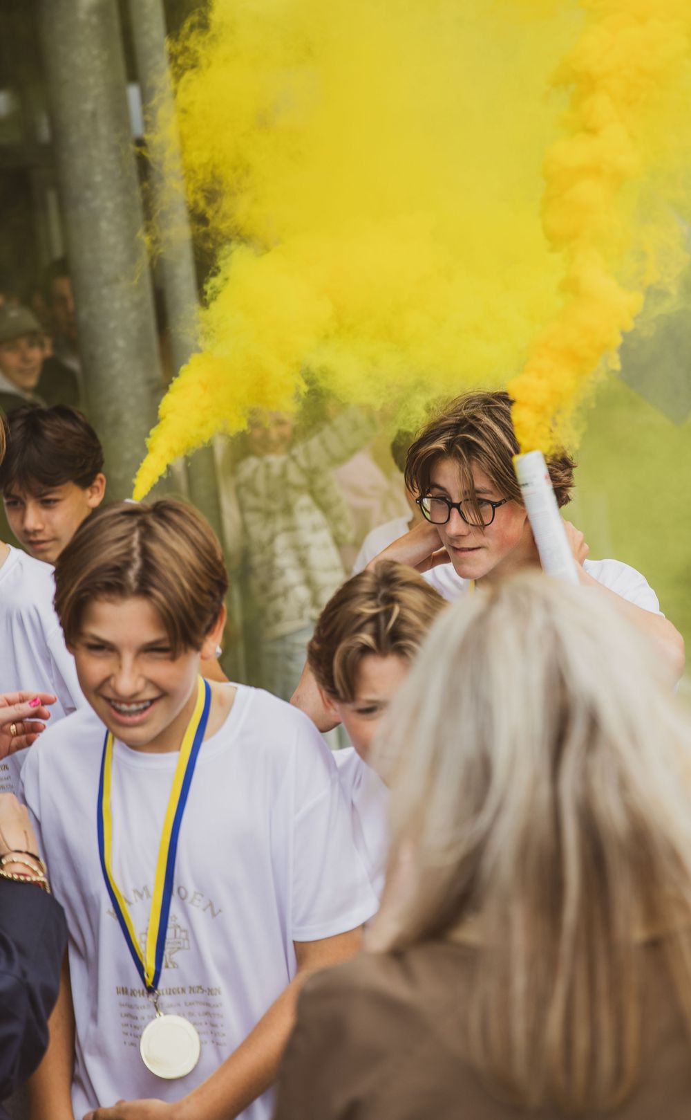 Students in white shirts celebrate with yellow smoke flare, one wearing a medal, at an outdoor school event.