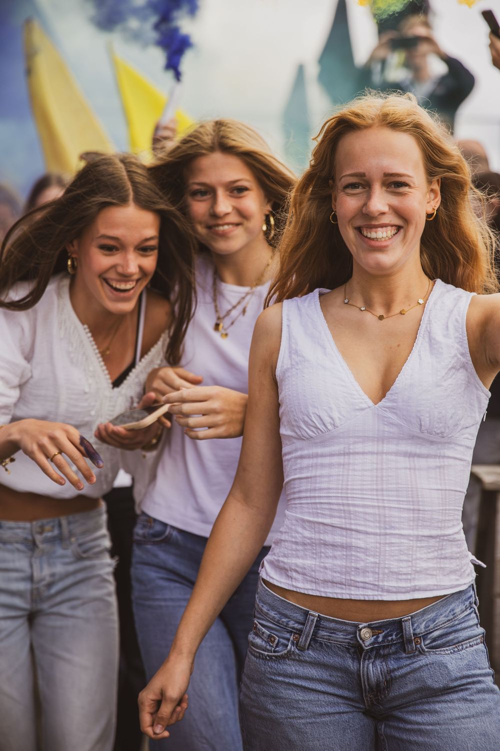 Three young women smiling and laughing together at an outdoor festival, wearing white tops and jeans.
