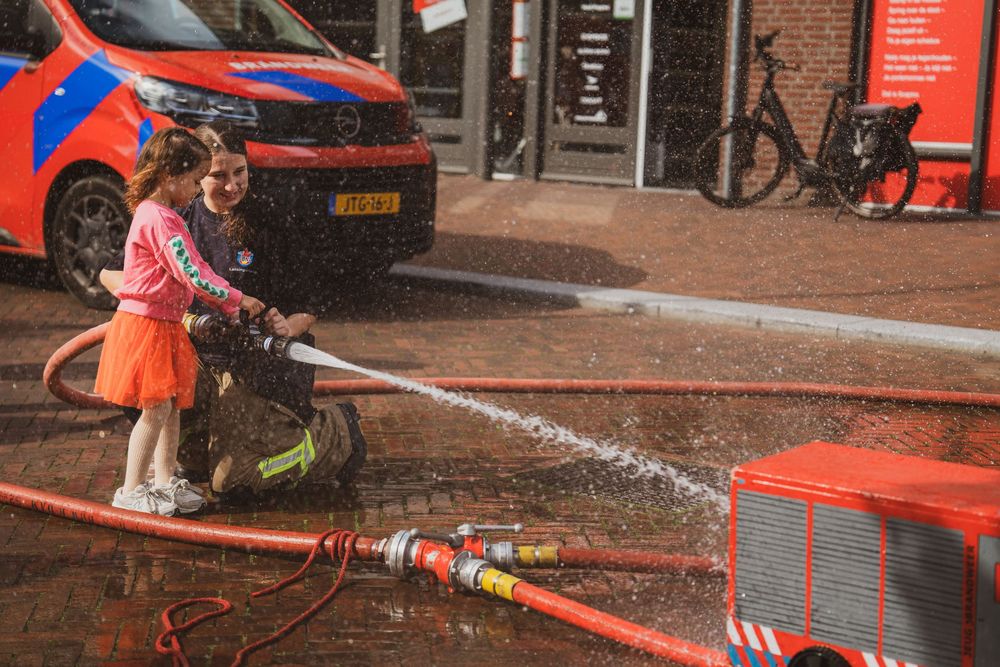 A firefighter helps a young girl operate a fire hose near a red fire truck and Dutch emergency vehicle.