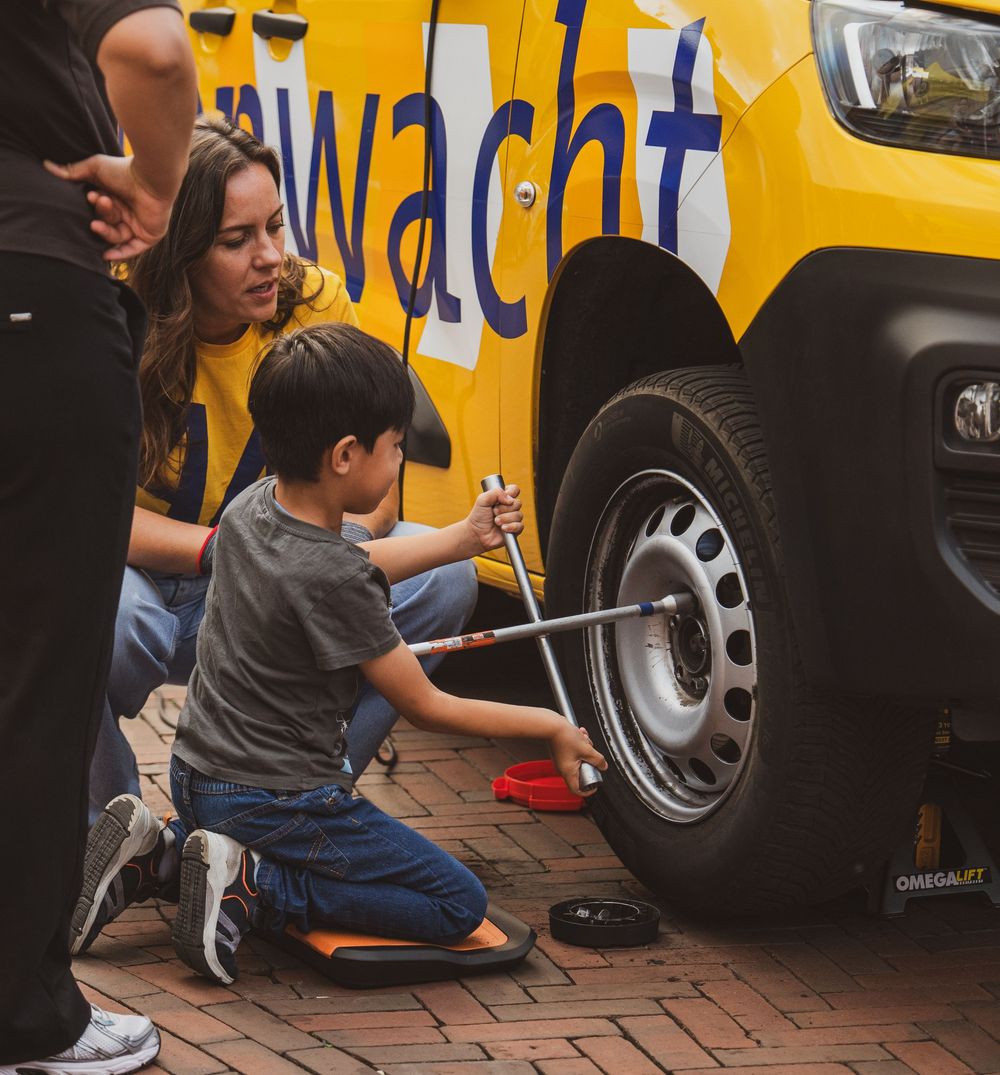 A young boy changes a tire on a yellow ANWB roadside assistance vehicle while a woman supervises on brick pavement.