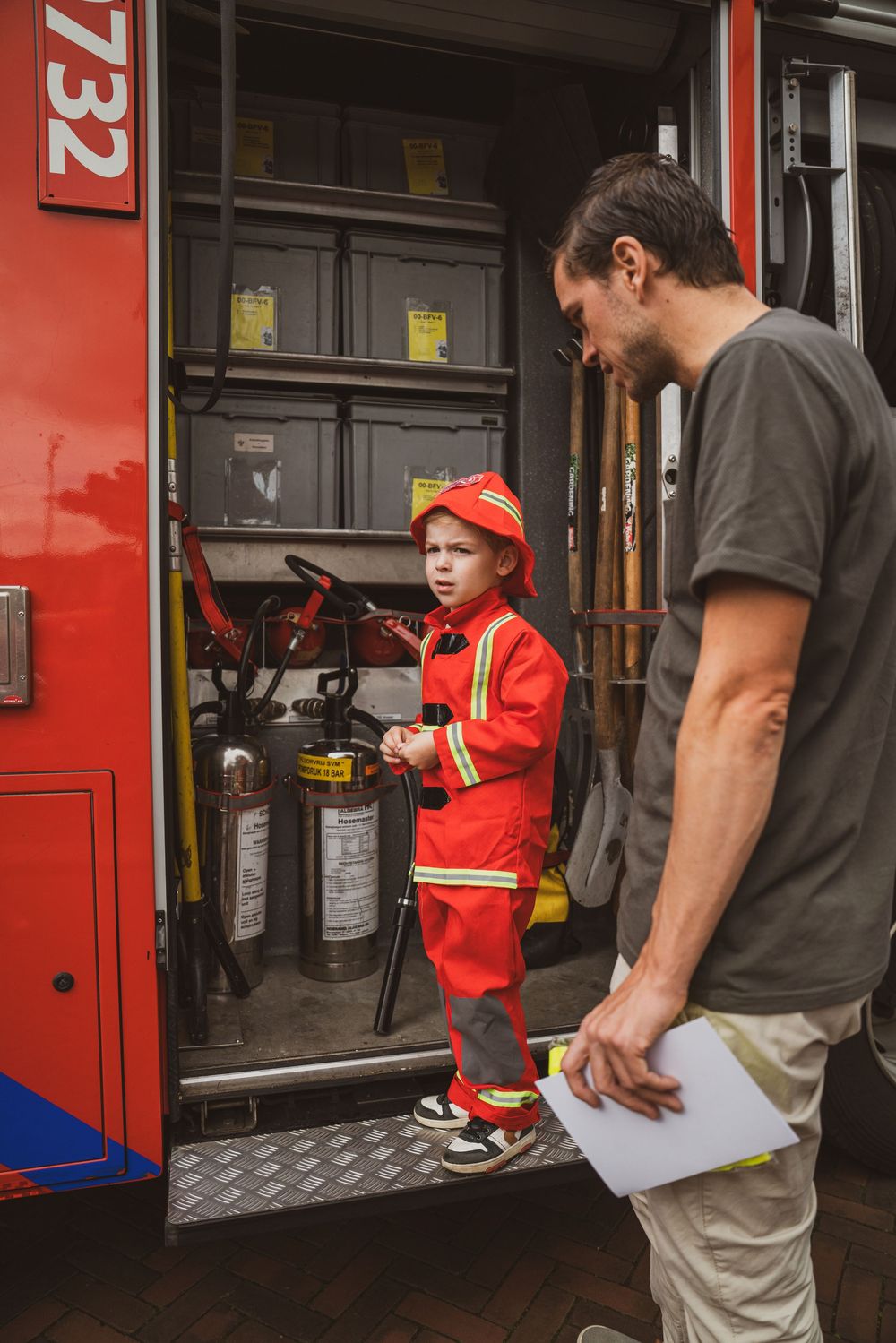 A young child in a red firefighter costume stands inside a fire truck while an adult man looks on attentively.