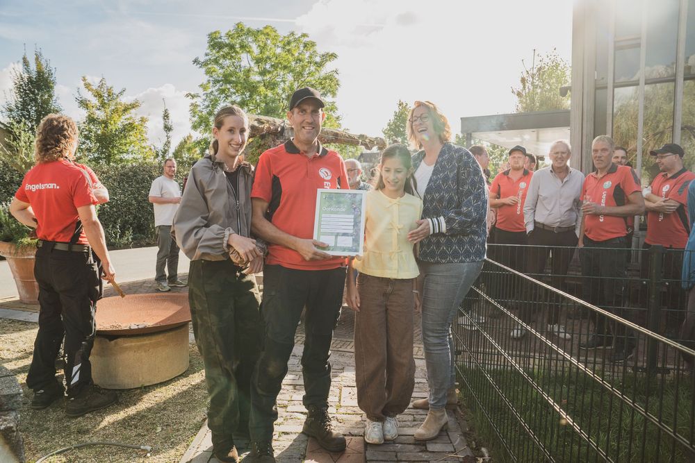 Group of people in red uniforms presenting a certificate outdoors at an animal enclosure on a sunny day.