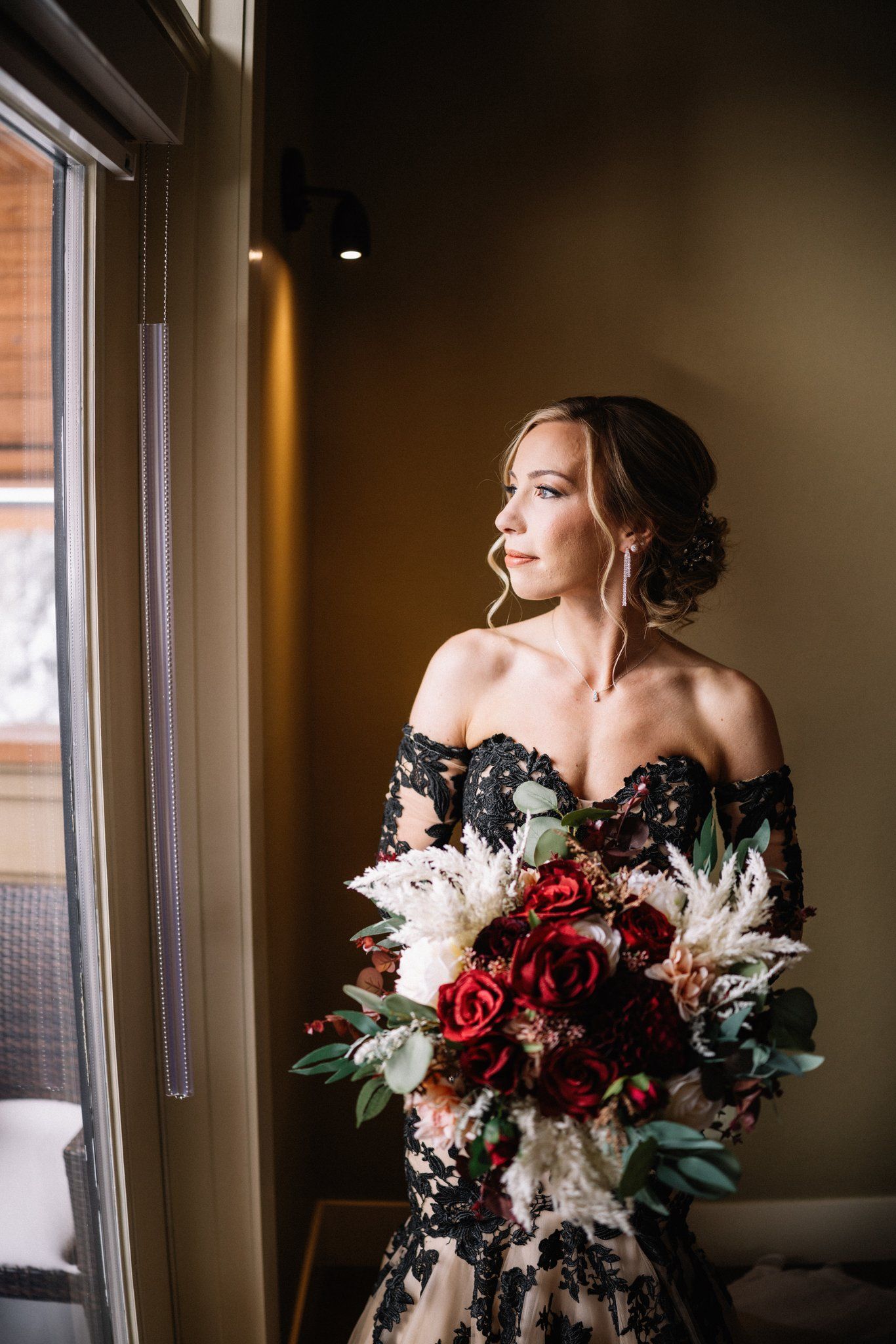 Bride in an off-the-shoulder black and ivory lace Sophia Tolli gown holding a red rose bouquet in window light