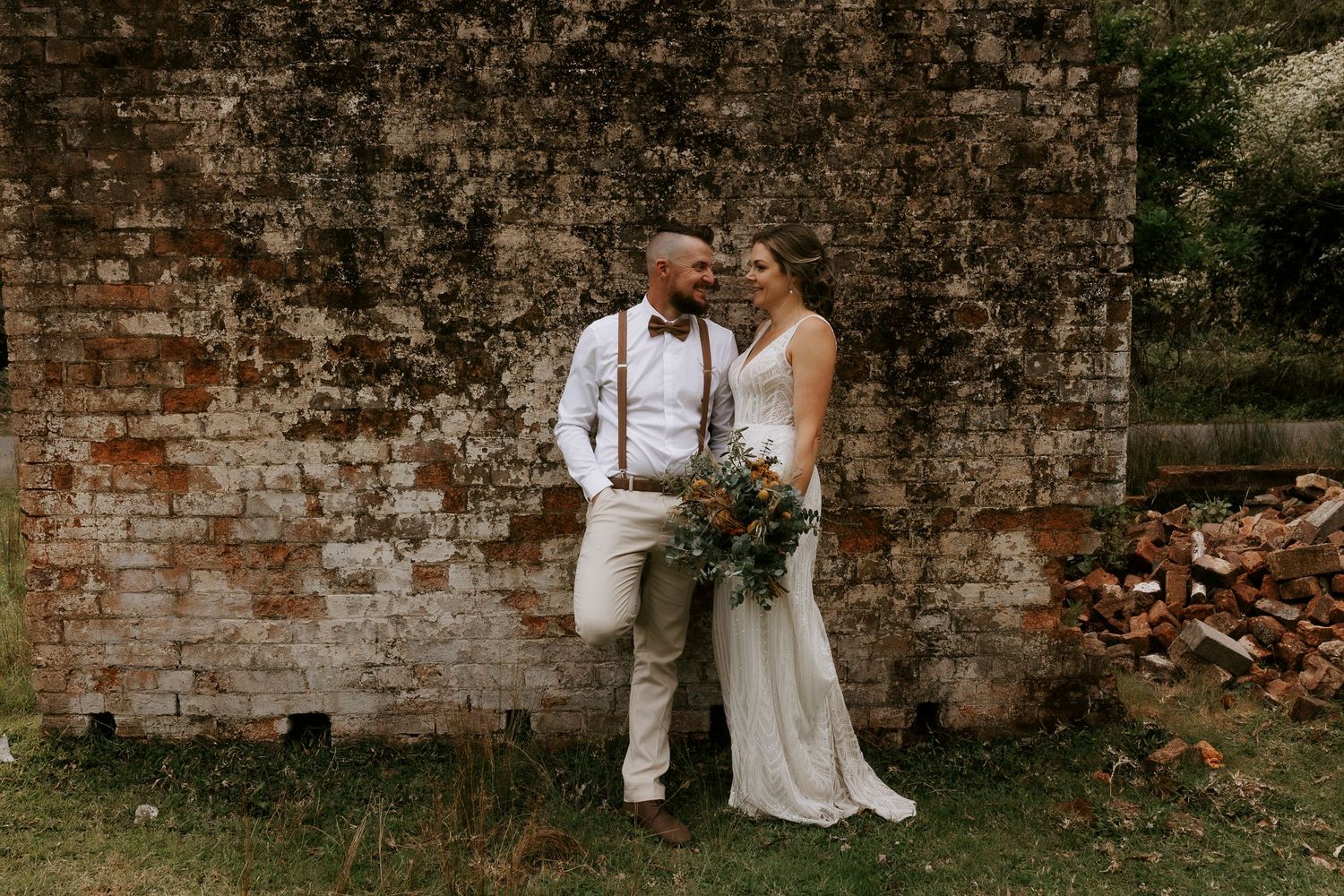 Wedding party stands in front of weathered brick wall in outdoor setting.