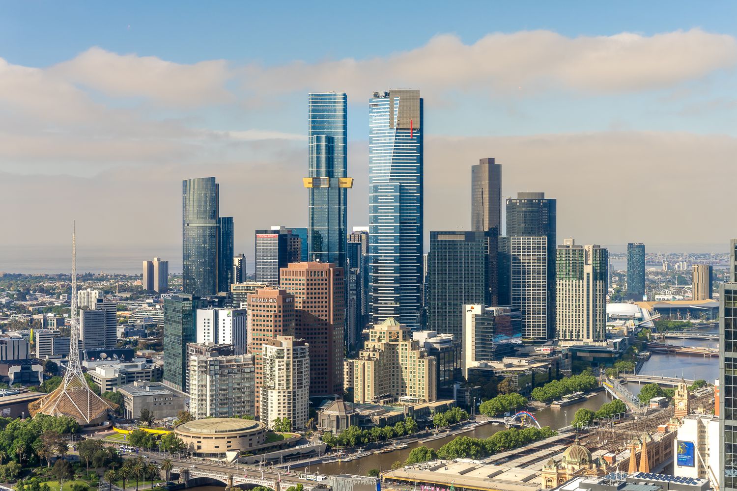 Aerial view of Melbourne CBD skyline with modern skyscrapers under a partly cloudy blue sky.