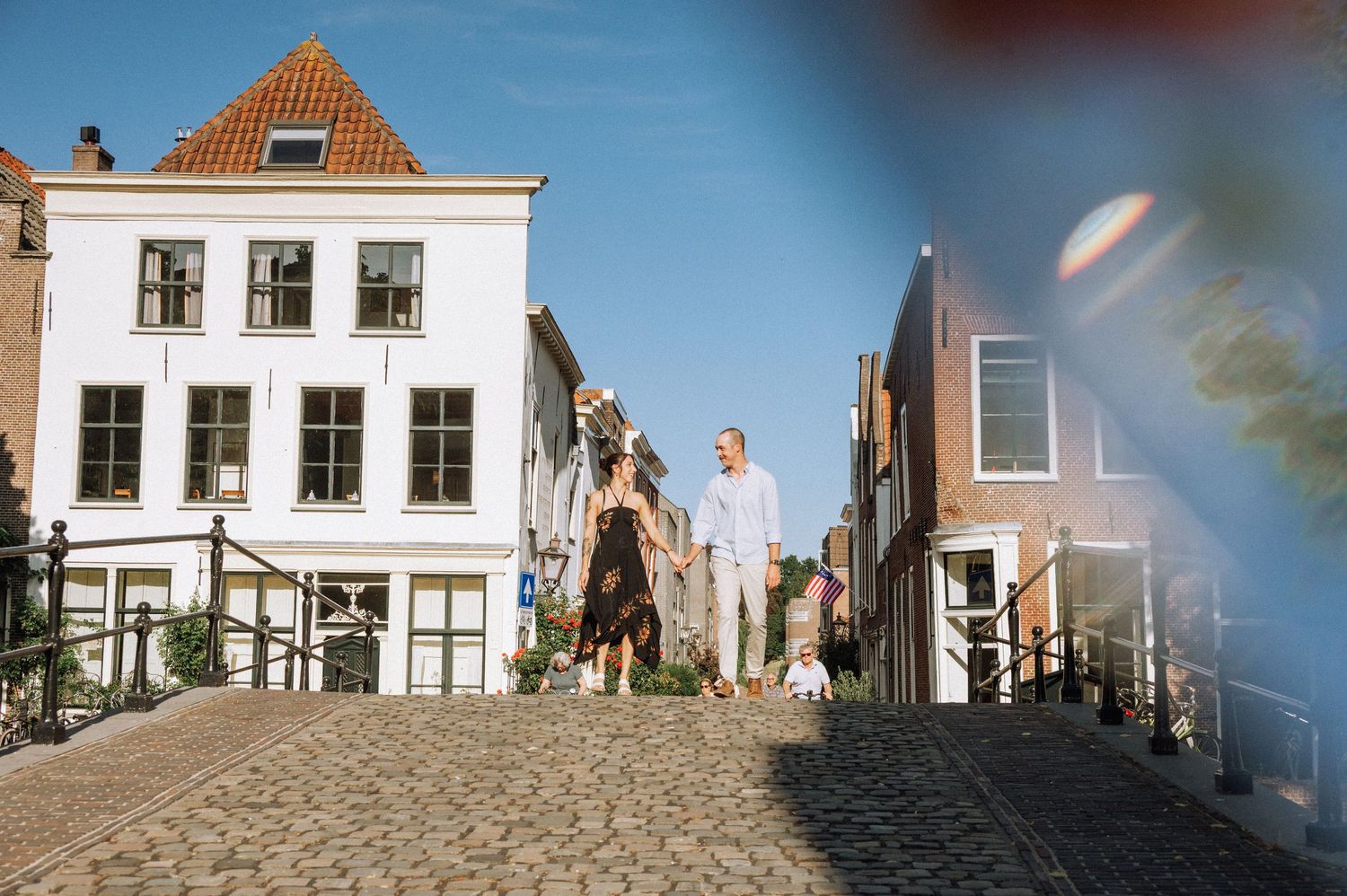 Couple walking hand in hand across a cobblestone bridge near historic white and red brick Dutch canal houses on a sunny day. Leiden
