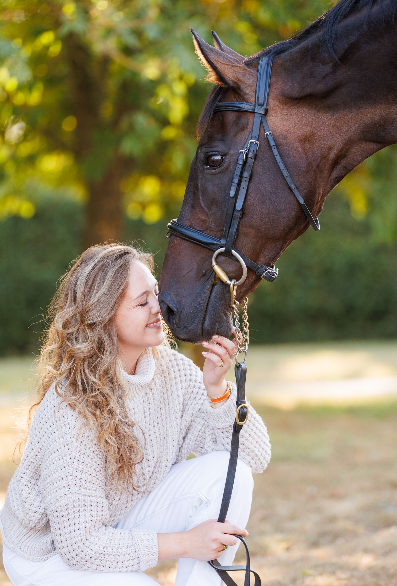 Woman walking two chestnut horses down a sunlit gravel path lined with green trees at a horse farm.